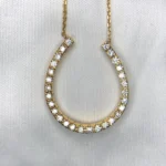 Macro photographs of a 14K yellow gold horseshoe diamond pendant necklace held against a white fabric backdrop, featuring 0.42 carats of natural round brilliant-cut diamonds prong-set along the horseshoe arch on a delicate gold chain.