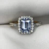 Close-up photograph of an 18K gold ring featuring a 1.53-carat emerald-cut pastel blue natural Ceylon sapphire centerpiece surrounded by a shimmering halo of 0.33-carat natural diamond accents against a soft white fabric background.