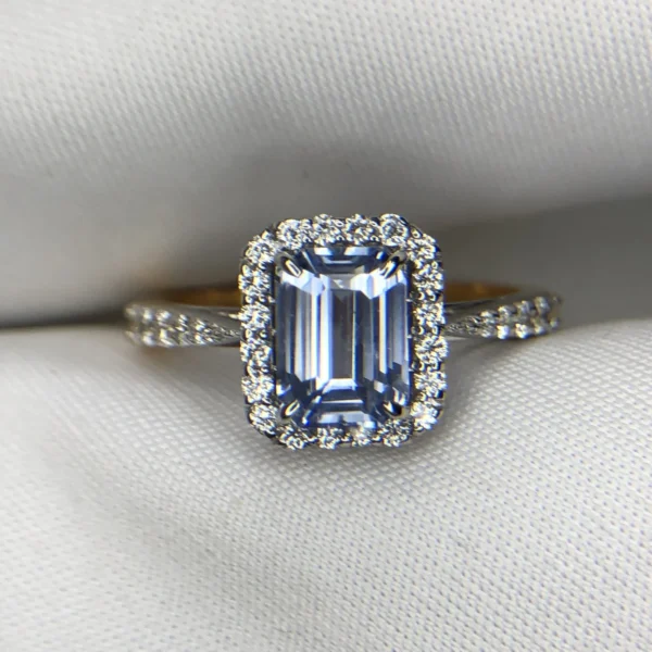 Close-up photograph of an 18K gold ring featuring a 1.53-carat emerald-cut pastel blue natural Ceylon sapphire centerpiece surrounded by a shimmering halo of 0.33-carat natural diamond accents against a soft white fabric background.