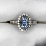 ​Close-up top-down photograph of an 18k gold diamond halo ring featuring a 1.72-carat natural pastel blue Ceylon sapphire (Neelam) accented by 0.33 carats of brilliant pavé diamonds, presented on a soft white fabric background.