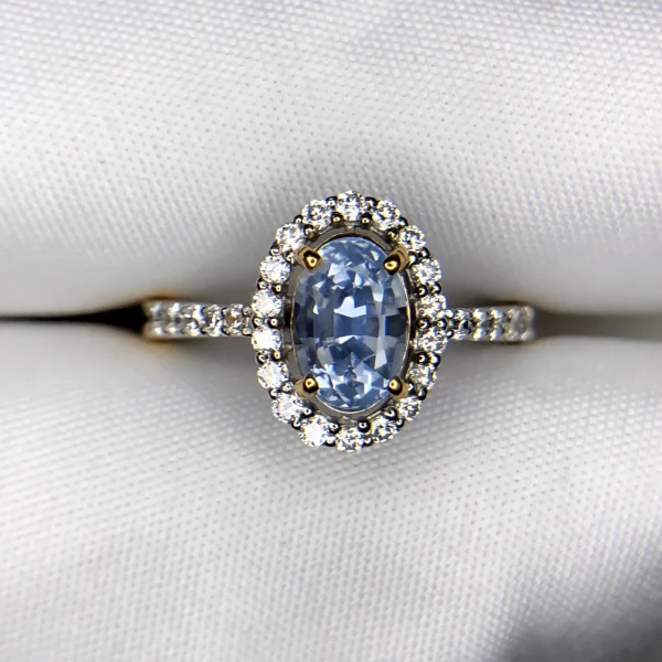 ​Close-up top-down photograph of an 18k gold diamond halo ring featuring a 1.72-carat natural pastel blue Ceylon sapphire (Neelam) accented by 0.33 carats of brilliant pavé diamonds, presented on a soft white fabric background.