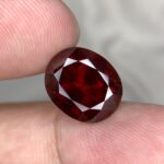 High-resolution macro photograph of a 9.81-carat natural Rhodolite Garnet held between fingers for scale reference, showcasing a masterfully cut oval silhouette, saturated deep crimson-red body color, crisp crown faceting, and exceptional gem-grade brilliance sourced from Madagascar.