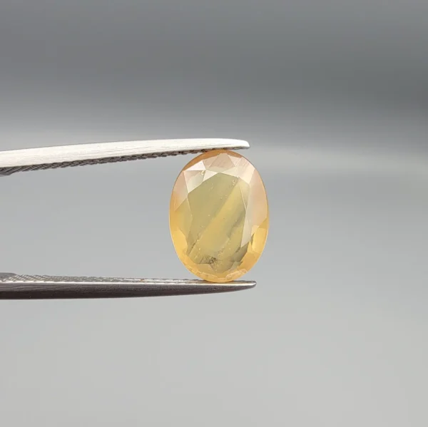 High-resolution macro photograph displaying a 3.78-carat natural Yellow Sapphire (Pukhraj) oval faceted gemstone held securely in gemological tweezers, featuring a warm golden-yellow body color, natural crystal inclusions, and brilliant facet architecture.