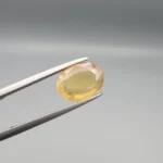 High-resolution macro photograph displaying a 3.78-carat natural Yellow Sapphire (Pukhraj) oval faceted gemstone held securely in gemological tweezers, featuring a warm golden-yellow body color, natural crystal inclusions, and brilliant facet architecture.