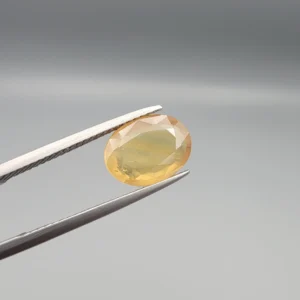High-resolution macro photograph displaying a 3.78-carat natural Yellow Sapphire (Pukhraj) oval faceted gemstone held securely in gemological tweezers, featuring a warm golden-yellow body color, natural crystal inclusions, and brilliant facet architecture.