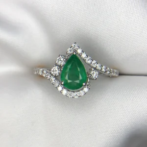 Close-up photograph of an 18K gold ring featuring a 1.73-carat pear-shaped vibrant green natural Zambian emerald centerpiece gracefully embraced by a winding wave of 0.56-carat natural diamond accents against a soft white fabric background.