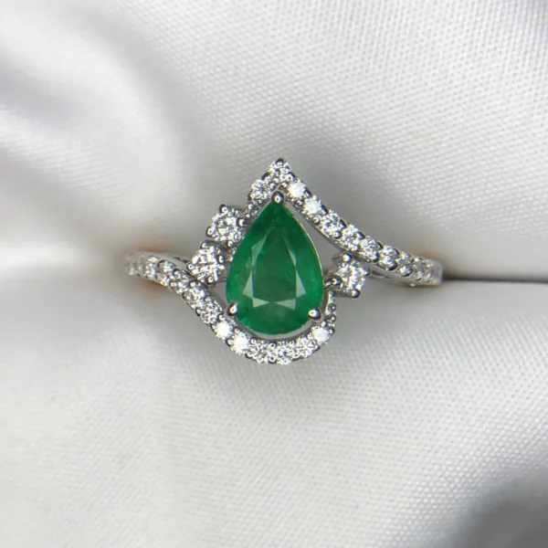 Close-up photograph of an 18K gold ring featuring a 1.73-carat pear-shaped vibrant green natural Zambian emerald centerpiece gracefully embraced by a winding wave of 0.56-carat natural diamond accents against a soft white fabric background.