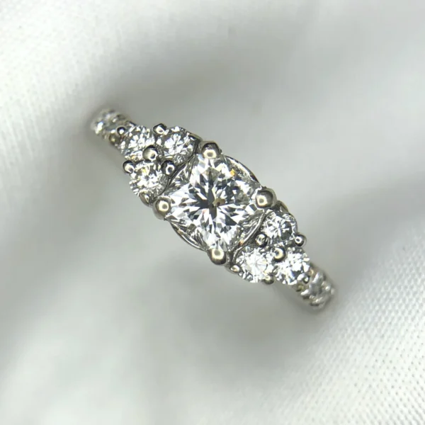 Multi-angle macro photographs of a 14K white gold 7-stone diamond engagement ring held against a white fabric backdrop, featuring a central natural cushion-cut diamond flanked by six graduated round brilliant-cut diamonds across the gallery band.