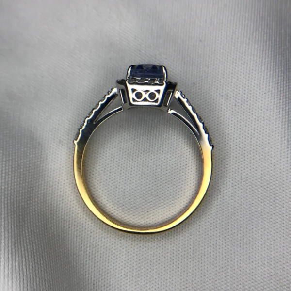 Close-up photograph of an 18K gold ring featuring a 1.53-carat emerald-cut pastel blue natural Ceylon sapphire centerpiece surrounded by a shimmering halo of 0.33-carat natural diamond accents against a soft white fabric background.