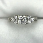 Multi-angle macro photographs of a 14K white gold 7-stone diamond engagement ring held against a white fabric backdrop, featuring a central natural cushion-cut diamond flanked by six graduated round brilliant-cut diamonds across the gallery band.