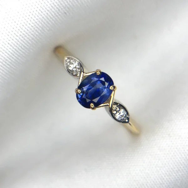 Macro photograph of a 1.91-gram 18K yellow gold fine ring set with a central 0.68-carat vibrant royal blue Ceylon sapphire flanked by sparkling diamond accents (0.04 CTW) on marquise-style shoulder settings against a white background.